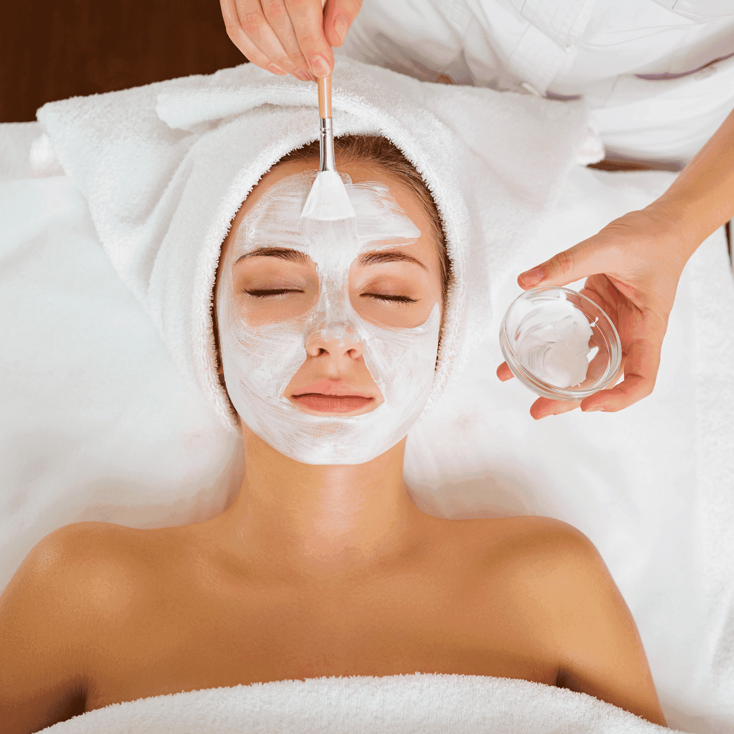 Woman receiving a facial treatment with a white mask applied using a brush, lying on a spa bed.