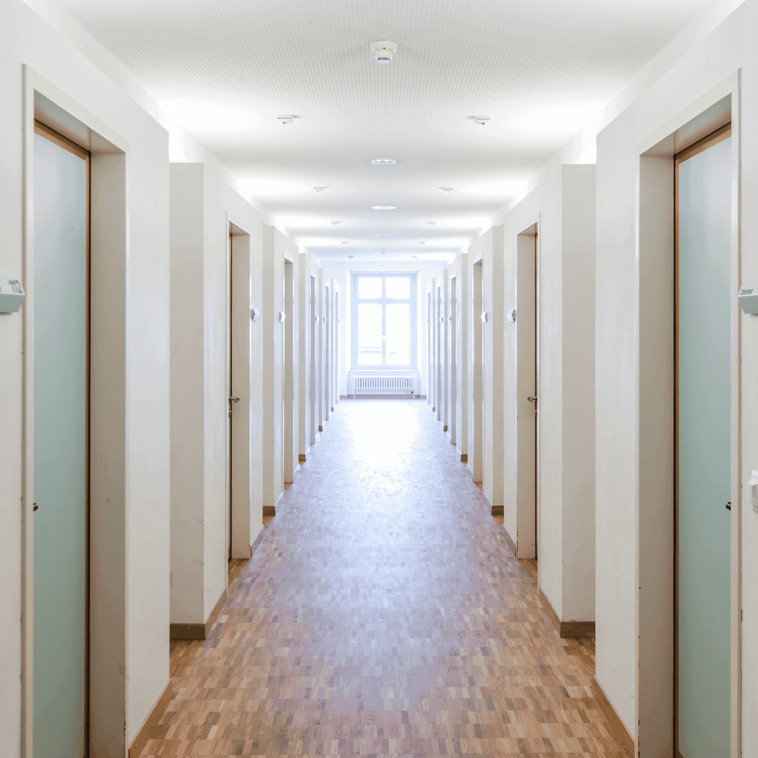 Bright, empty corridor with white walls, wooden floor, and multiple doors leading to a window.